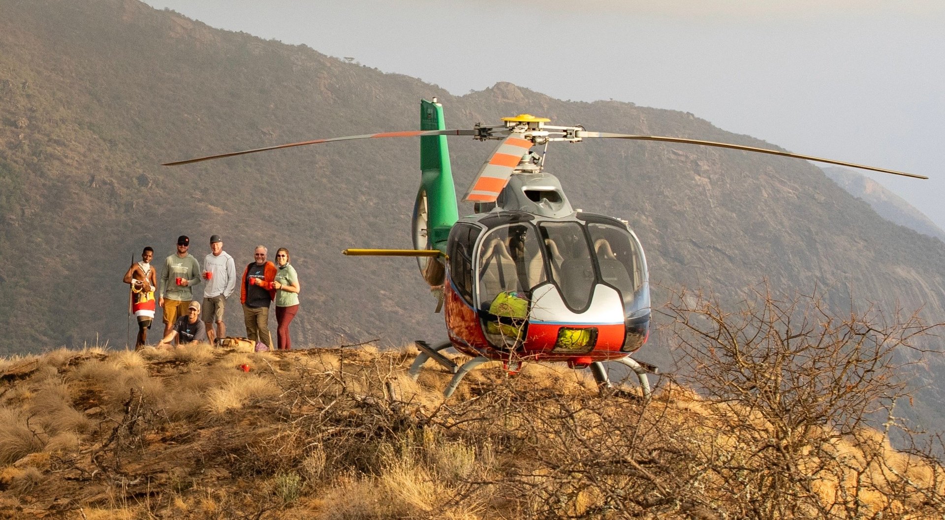 Green and yellow helicopter on rocky hillside with rescue team standing nearby, mountains in background